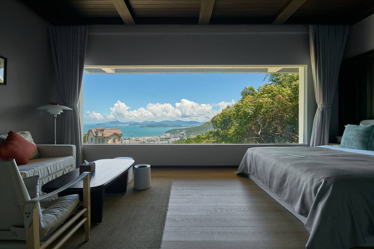 A bedroom with a large window, a bed, a sofa, a small table, and a chair.