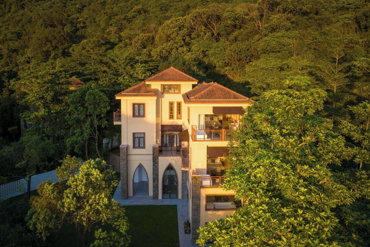 A large, multi-story house surrounded by dense forest, featuring a prominent entrance with a covered porch and multiple balconies.