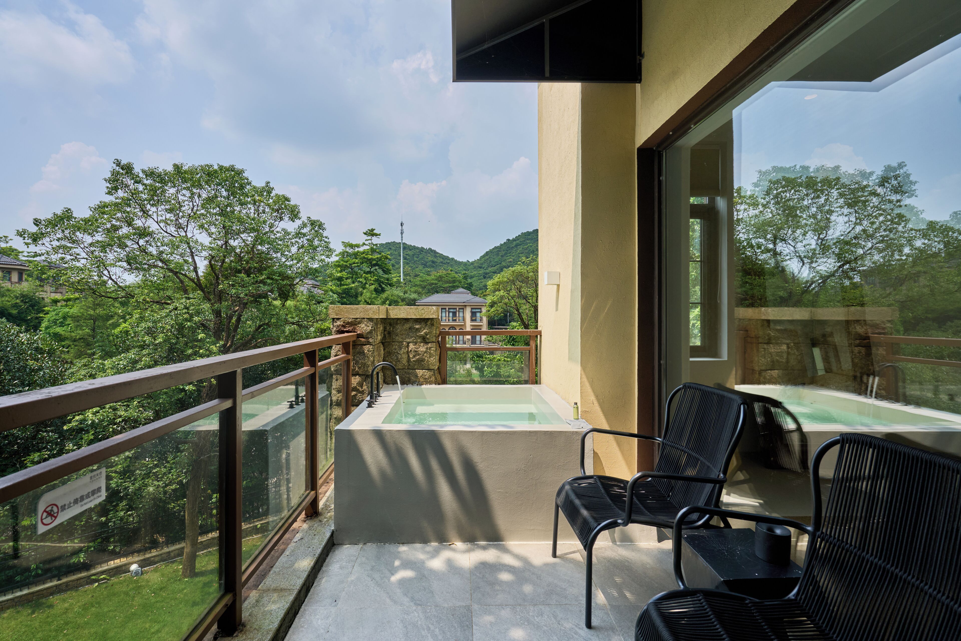 A balcony with a hot tub, two black chairs, and a view of a lush green landscape with trees and a distant building.