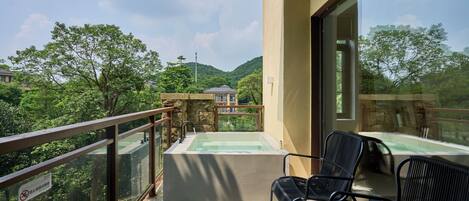 A balcony with a hot tub, two black chairs, and a view of a lush green landscape with trees and a distant building.