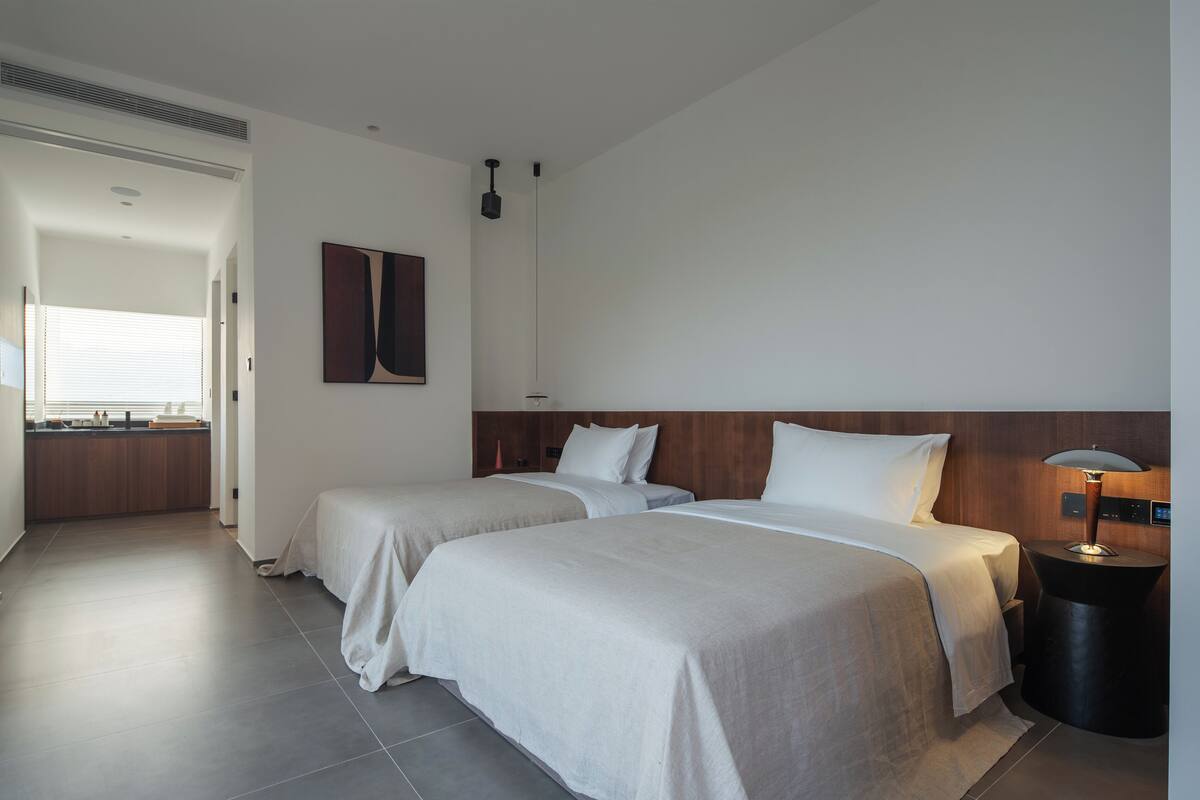 A hotel room with two beds, a bedside table with a lamp, a wall-mounted artwork, and a view of a kitchen area.