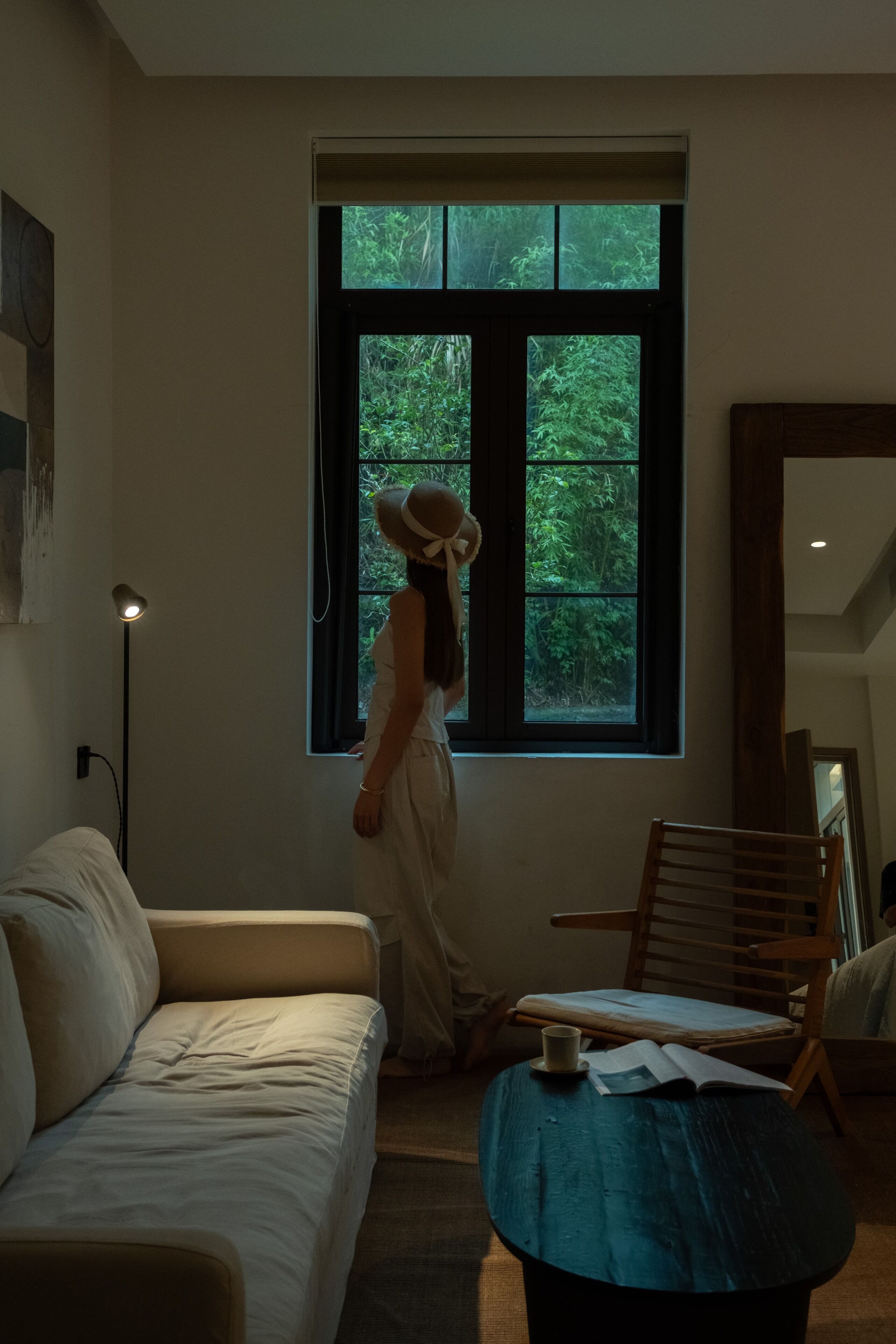 A room with a large window, a sofa, a wooden chair, and a small table with a book and a cup.