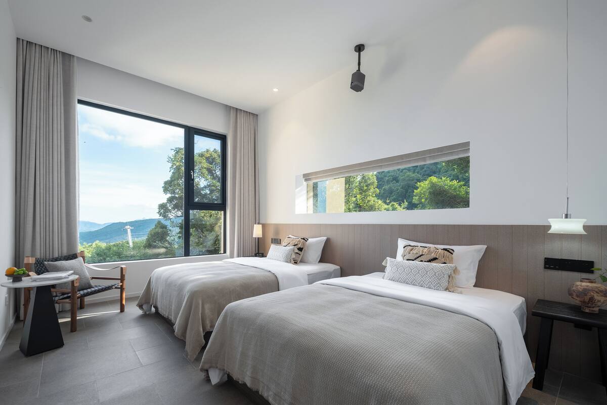 A modern hotel room with two beds, a small table with a chair, a view of the outdoors, and a large window.