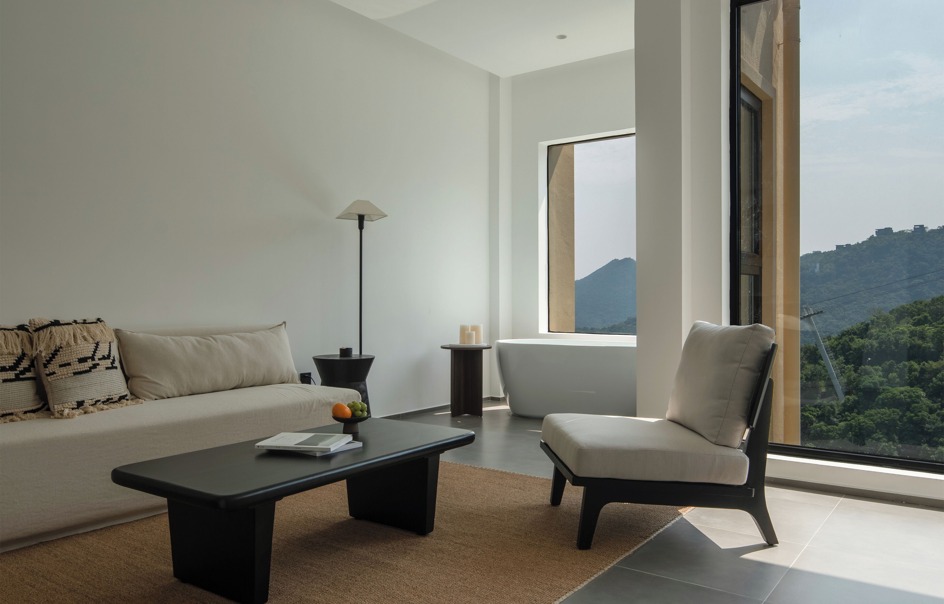 A modern living room with a sofa, a chair, a coffee table, and a lamp. There is a large window with a view of a mountain and greenery.