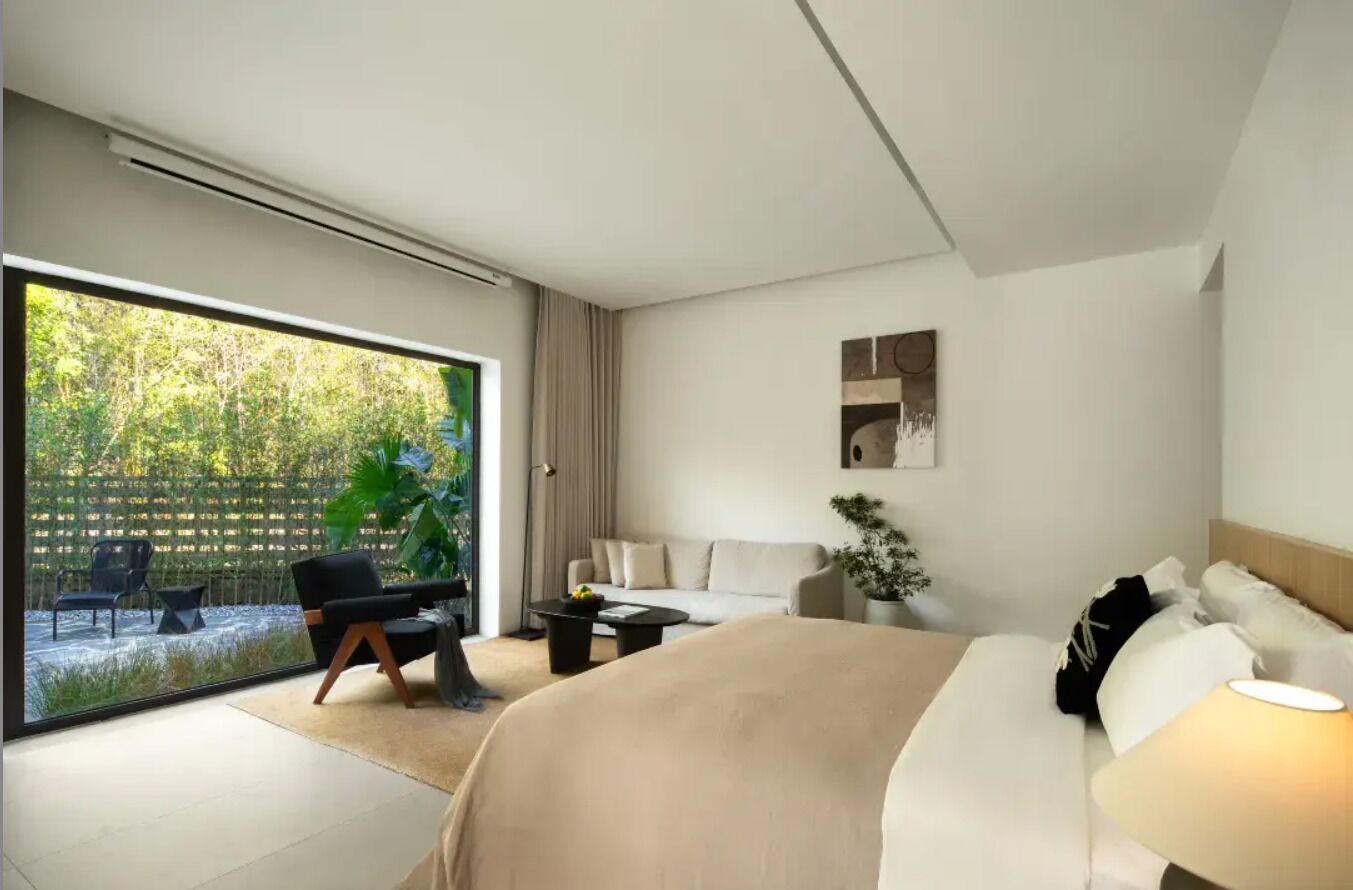 Ein modernes Hotelzimmer mit einer großen Glasschiebetür, die zu einem Garten führt, einem Bett mit beiger Bettdecke, einem kleinen runden Tisch, einer Couch und einer Topfpflanze.