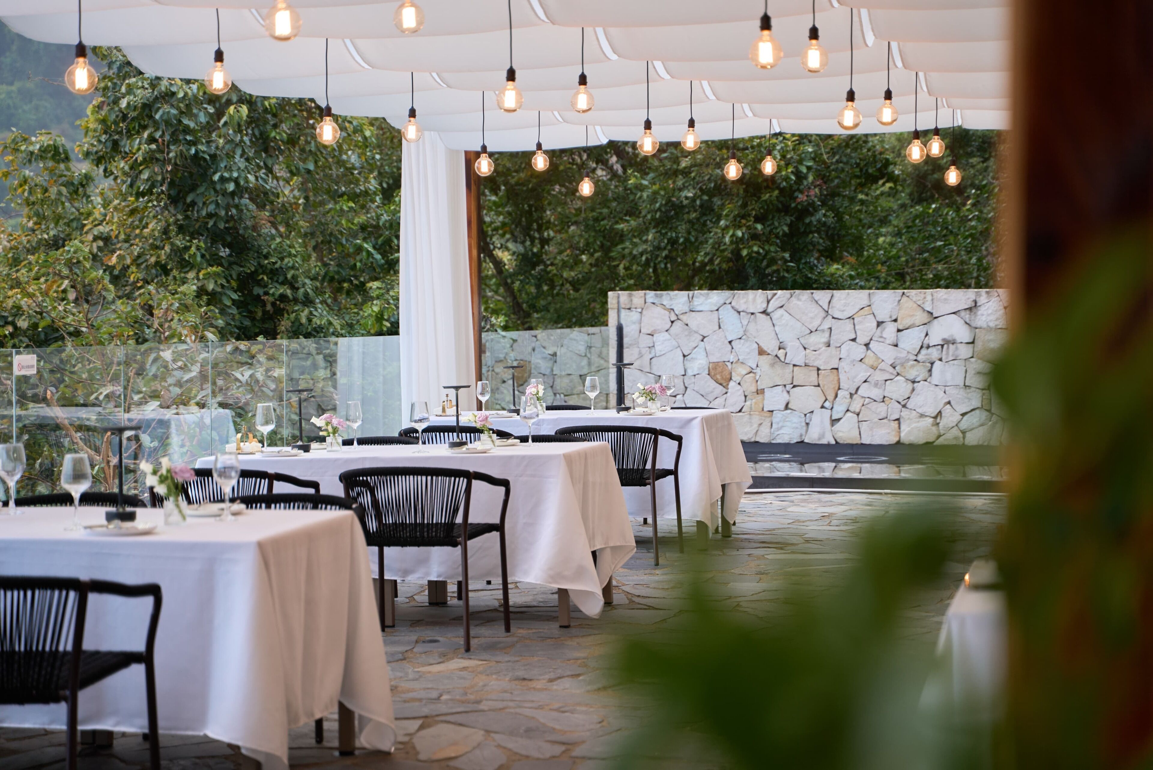 An outdoor dining area with tables set for a meal, surrounded by a stone wall and greenery.