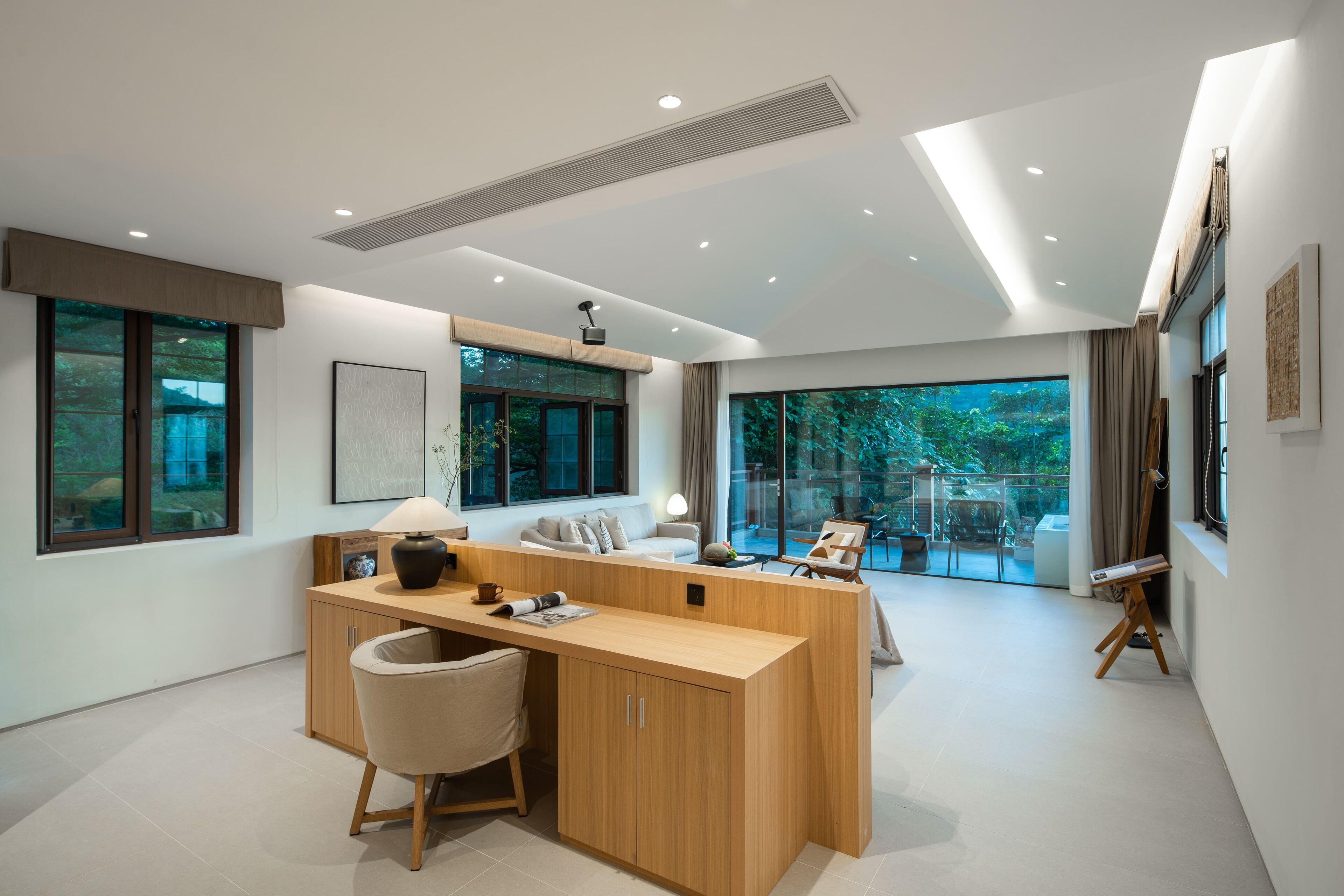A modern, spacious living area with a wooden desk, comfortable seating, and large windows offering a view of greenery.