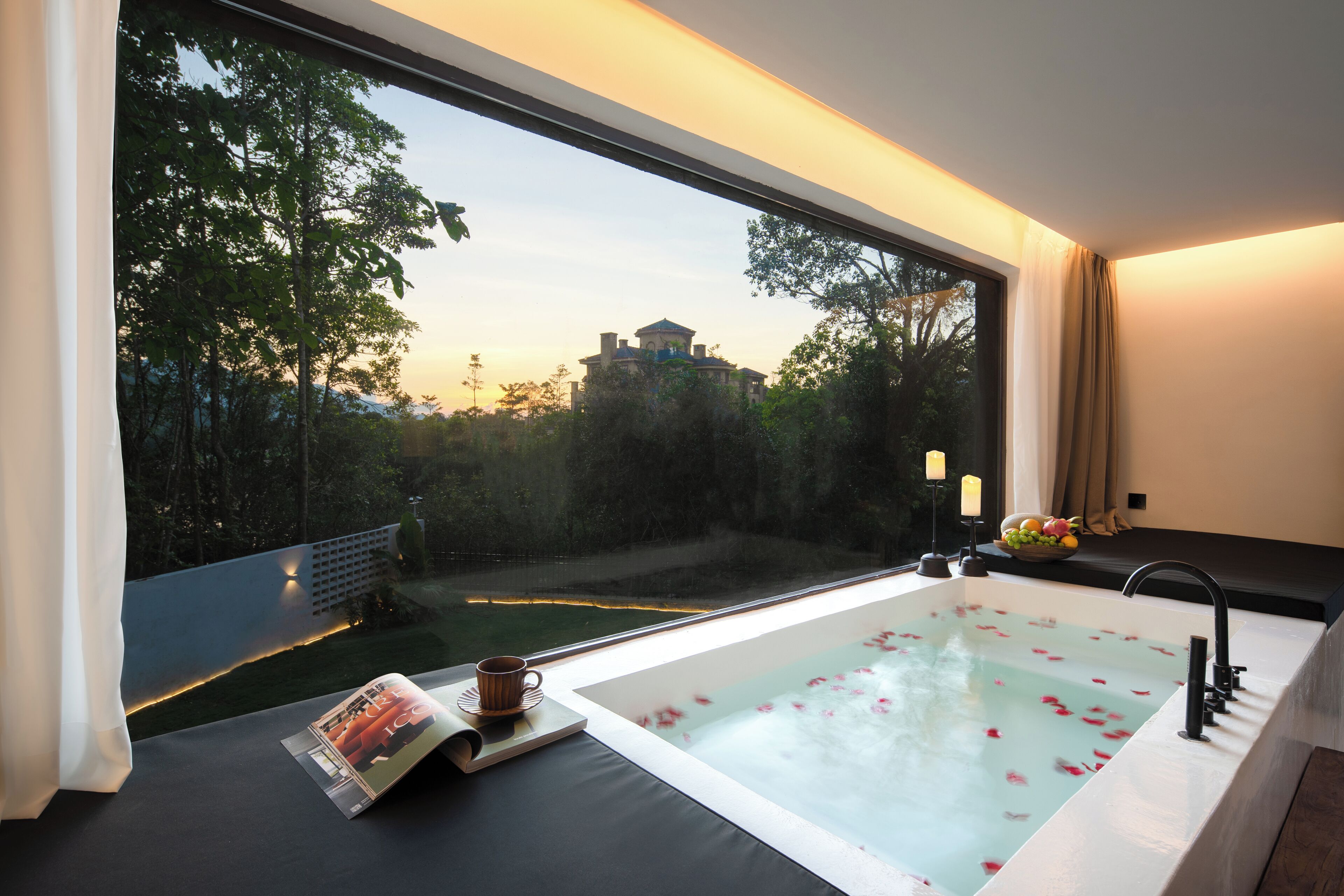 Un baño moderno con una bañera grande, una revista, una taza y vistas a un edificio a través de un ventanal amplio.