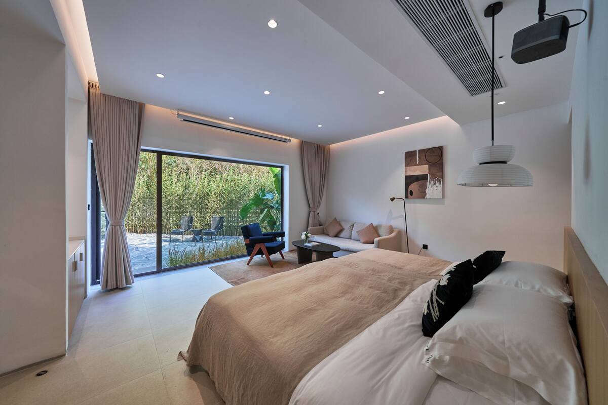 A modern bedroom with a large bed, a sofa, a coffee table, and a view of the outdoors.