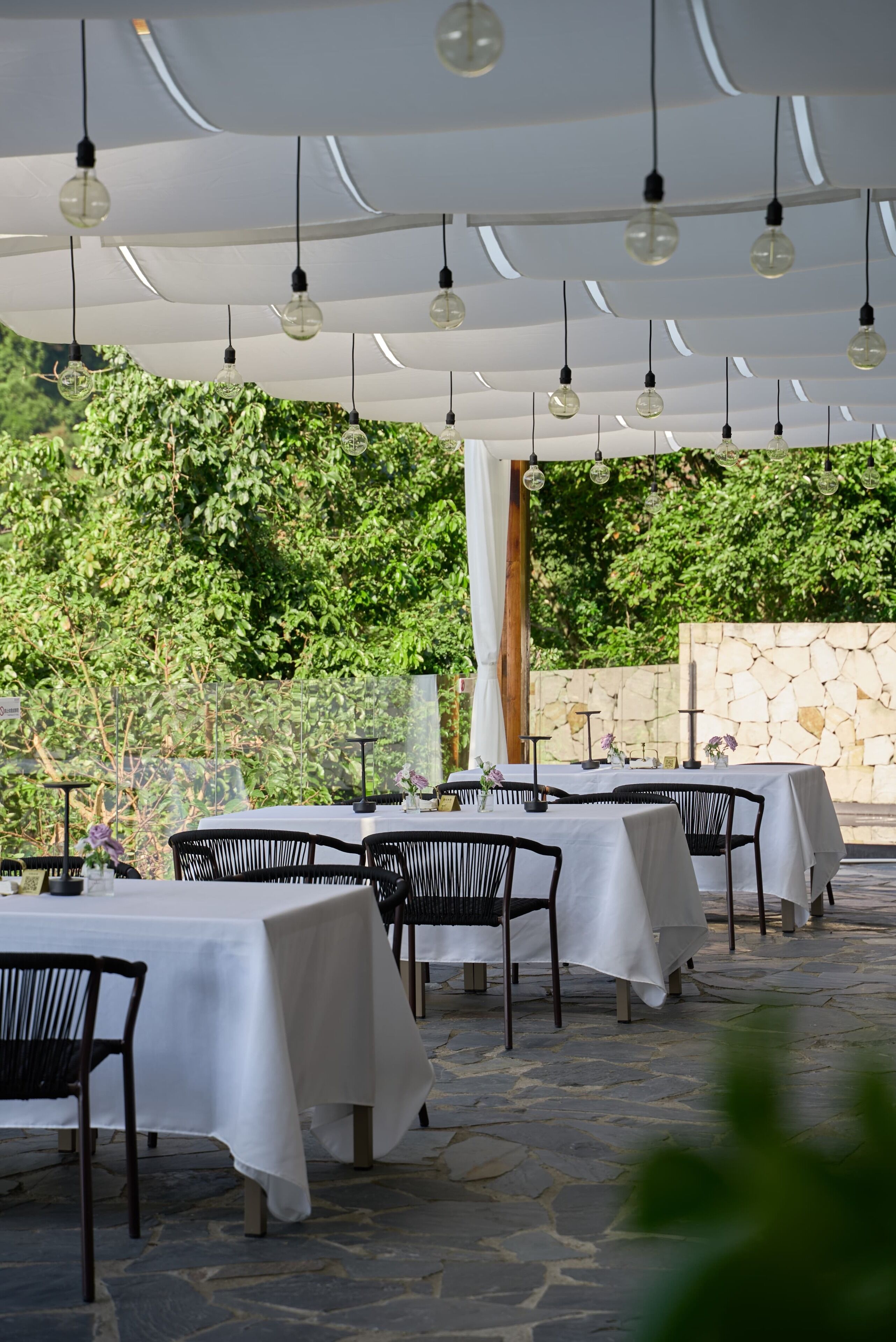 An outdoor dining area with tables and chairs, covered by a white canopy, and hanging light fixtures.