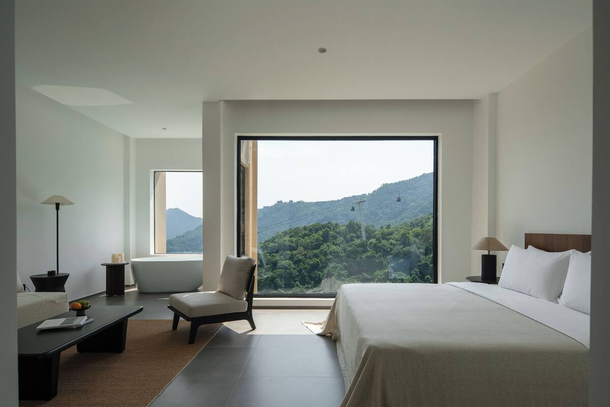 A modern hotel room with a large bed, a chair, a desk, and a view of a mountainous landscape.