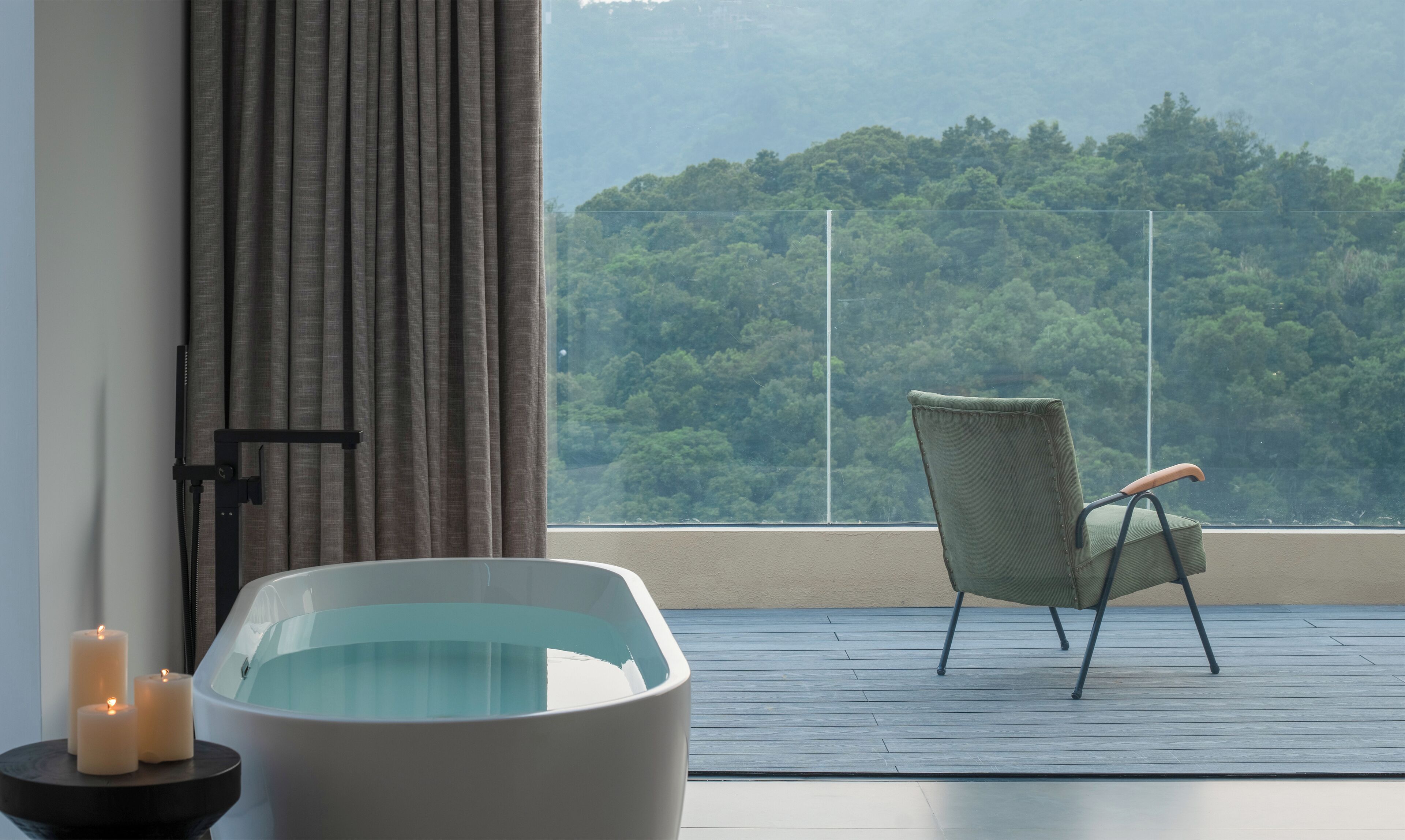 A room with a bathtub, a chair, and a view of a forested hill.