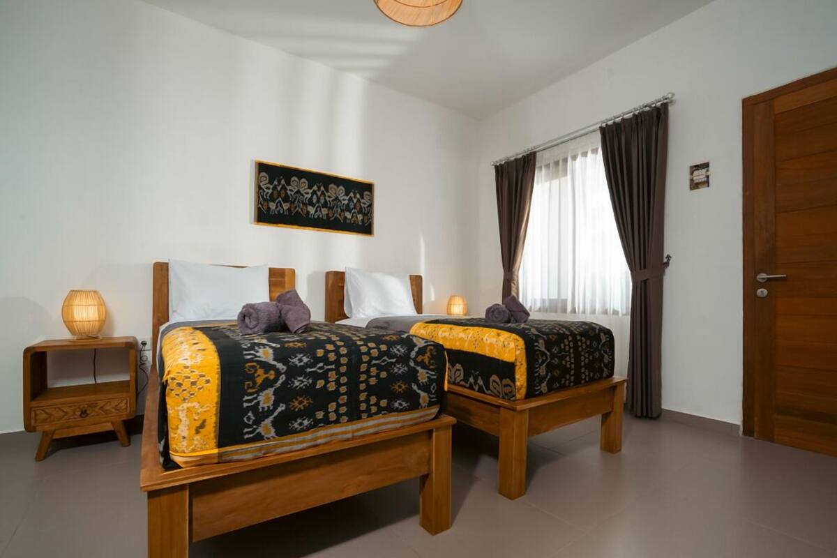 A room with two wooden beds, each with patterned bedding, a bedside table with a lamp, and a wall-mounted artwork.