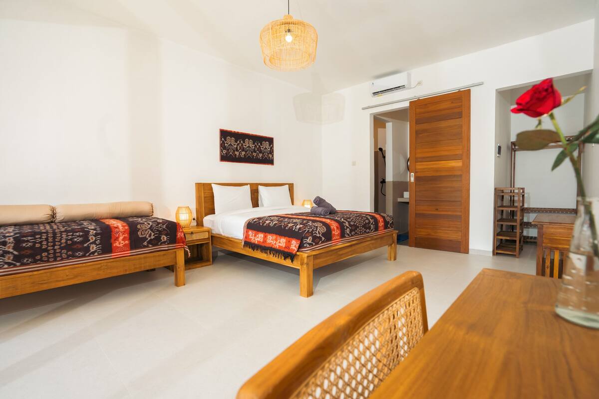 A bedroom with a wooden bed, a wooden table, and a chair. There is a wooden door and a wall-mounted air conditioning unit.