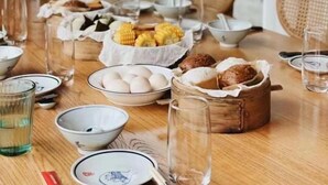 Een eettafel gedekt met diverse gerechten, waaronder maïs, eieren en brood, in een kamer met uitzicht op groen.