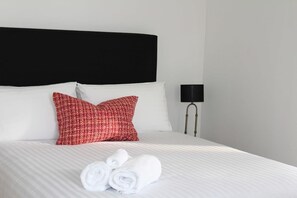 Ein ordentlich bezogenes Bett mit weißen Leinen, zwei Kissen (eins rot, eins weiß) und zusammengerollten Handtüchern.