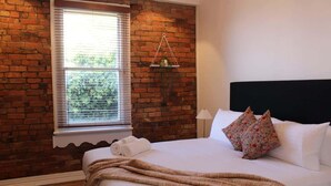 A hotel room with a brick wall, a window with blinds, a bed with white linens, and a hanging plant. - Forest Rd Haven with Tamar River Views (Trevallyn)