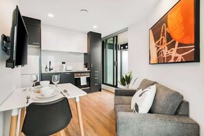 Une cuisine moderne avec des meubles blancs, une table à manger dressée pour deux personnes et un canapé gris dans un espace de vie agrémenté d’un grand tableau abstrait.