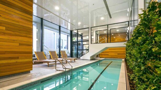 A modern indoor pool with clear glass walls, wooden paneling, and a staircase leading to an upper level. - Prime Location 1BR vs Aricon&Pool beside Skytower (Auckland)