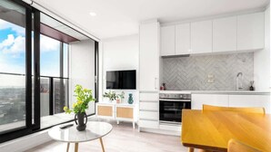 A modern kitchen with white cabinets, a herringbone backsplash, and a built-in oven. There's a dining area with a wooden table and chairs, and a small white table with a vase of flowers. - Incredible View Aircon 1B Studio Auckland Central (Auckland)