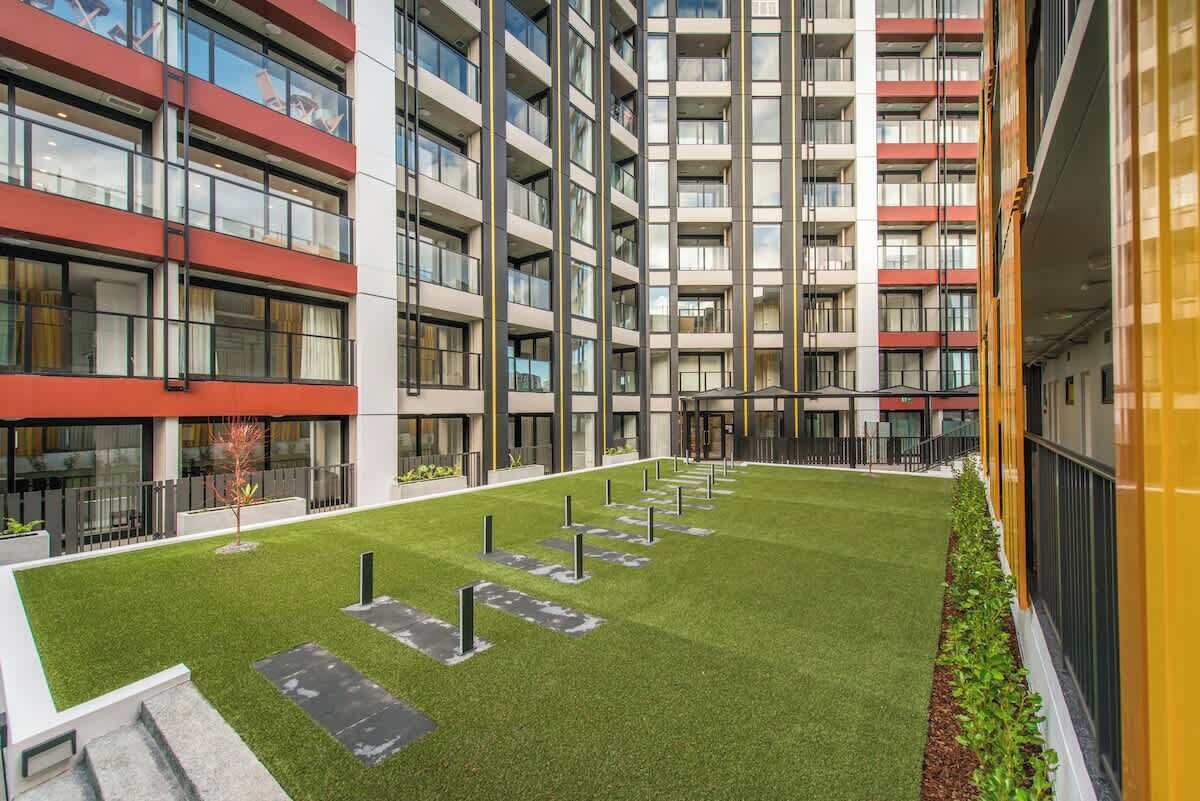 Un moderno edificio de apartamentos con varios balcones, un patio central con césped artificial y un edificio amarillo a la derecha.