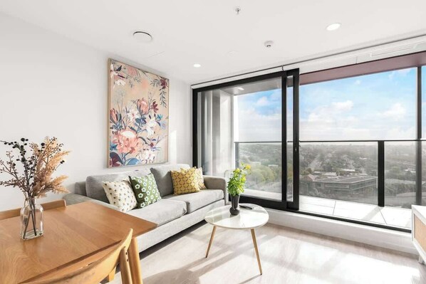 A modern living room with a large window, a sofa with patterned cushions, a dining table with chairs, and a decorative vase with dried flowers. - Incredible View Aircon 1B Studio Auckland Central (Auckland)