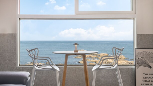 Una habitación con un ventanal grande con vistas a una costa rocosa y al mar. Hay una mesa redonda con dos sillas y un farol encima.