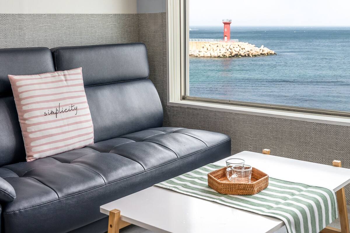 Una sala de estar moderna con un sofá de cuero, un cojín a rayas, una mesita con un vaso y una cesta, y vista al mar y un faro.