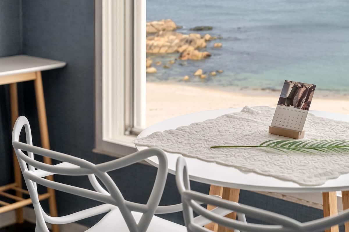 Una mesa redonda con un calendario y una hoja sobre un mantel de encaje, con vista a una playa con rocas y agua.