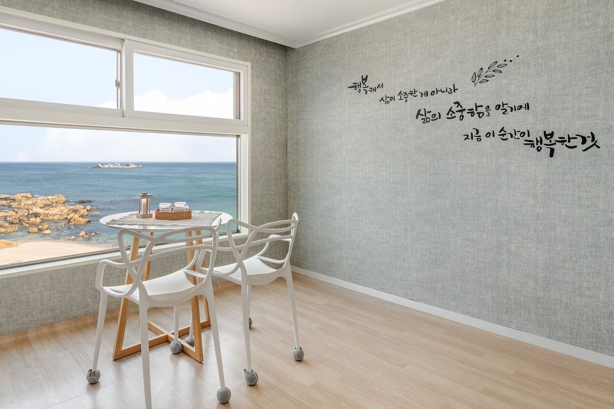 Una habitación con un ventanal grande que da a una playa rocosa y al mar. Hay una mesa redonda con sillas y una mesita con un farolillo y libros. Las paredes tienen un papel tapiz texturizado con texto coreano.
