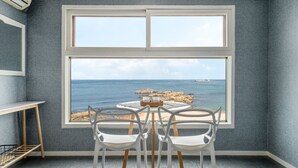 Una habitación con un ventanal grande que da a una costa rocosa y al mar. Hay una mesa redonda con cuatro sillas blancas y una pequeña cesta encima.