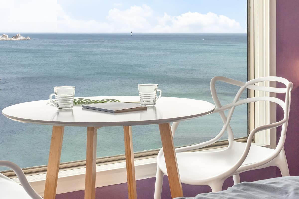 Una habitación con una mesa redonda y dos sillas blancas, una ventana con vista al mar y un faro, y una pared morada.