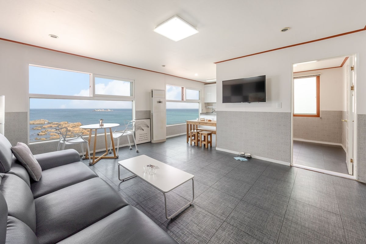 Una sala de estar moderna con un sofá gris, una mesita y un televisor montado en la pared. Una ventana grande ofrece vistas al mar.