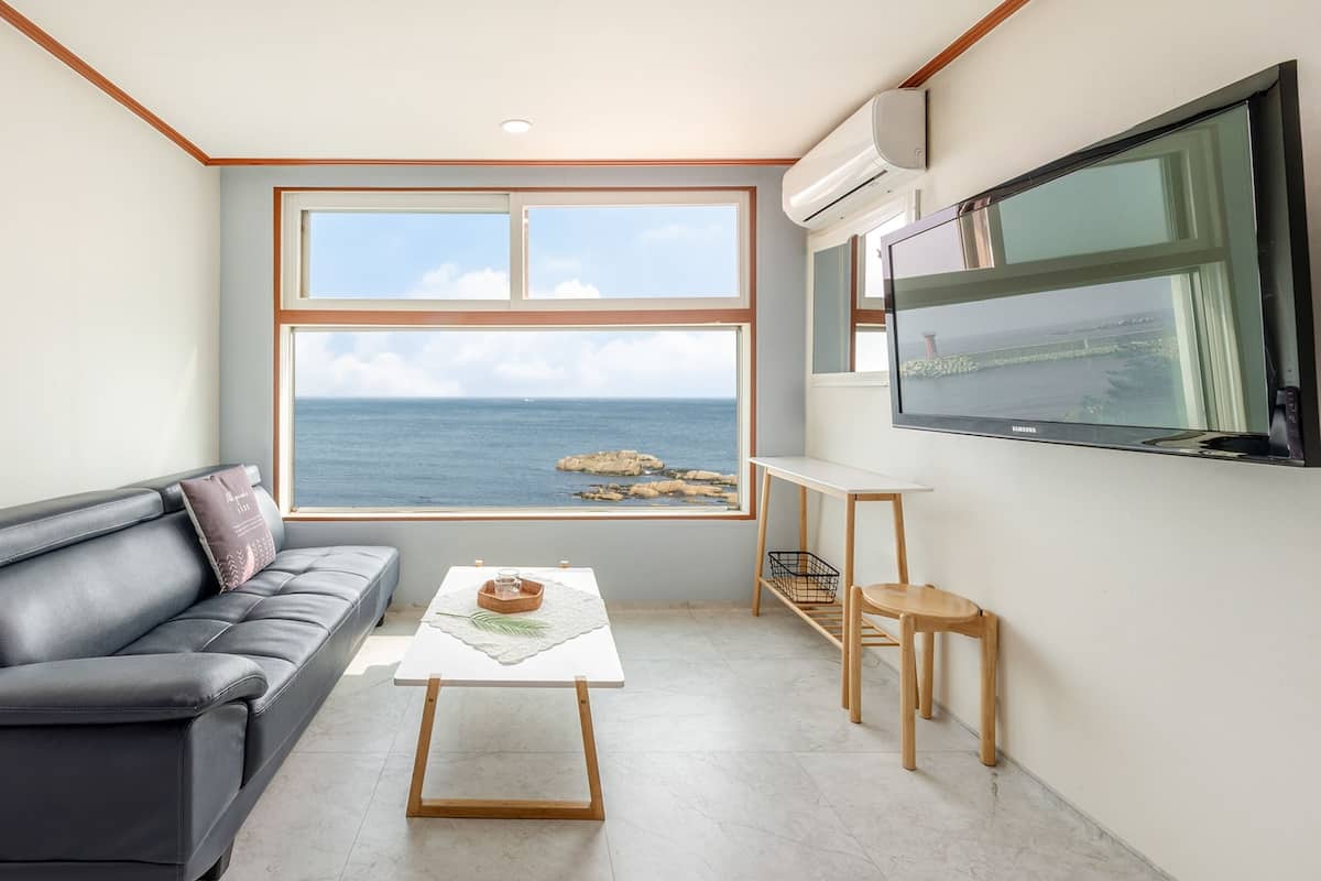 Un salón moderno con un sofá de cuero negro, una mesita blanca pequeña y un televisor montado en la pared. Un ventanal grande ofrece vistas al mar y a las rocas.