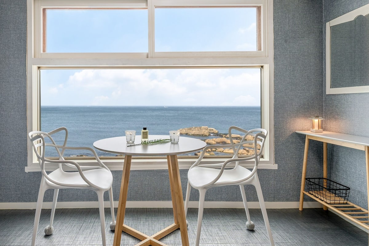 Una habitación con un ventanal grande que da al mar, dos sillas blancas, una mesa redonda y un estante pequeño con una lámpara.