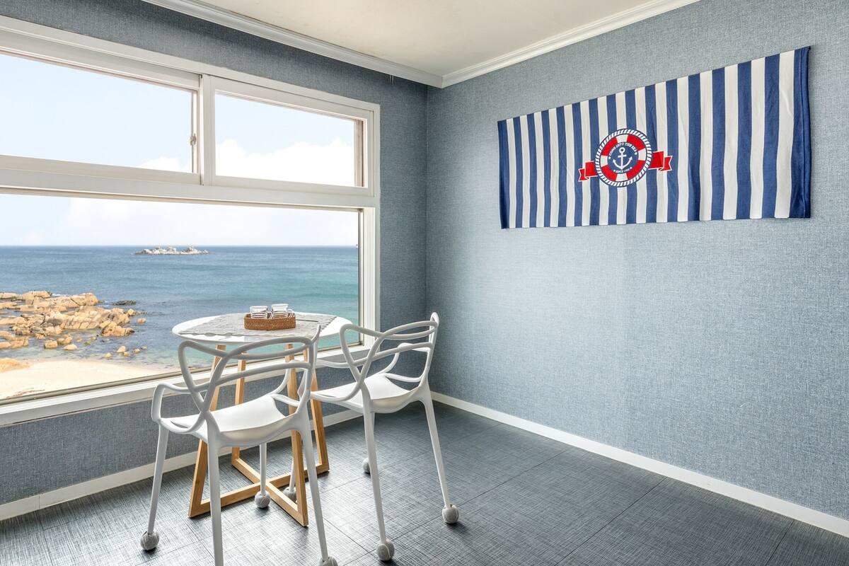 Una habitación con un gran ventanal con vistas a una costa rocosa y al mar. Hay una mesa redonda con sillas y un adorno a rayas colgado en la pared.