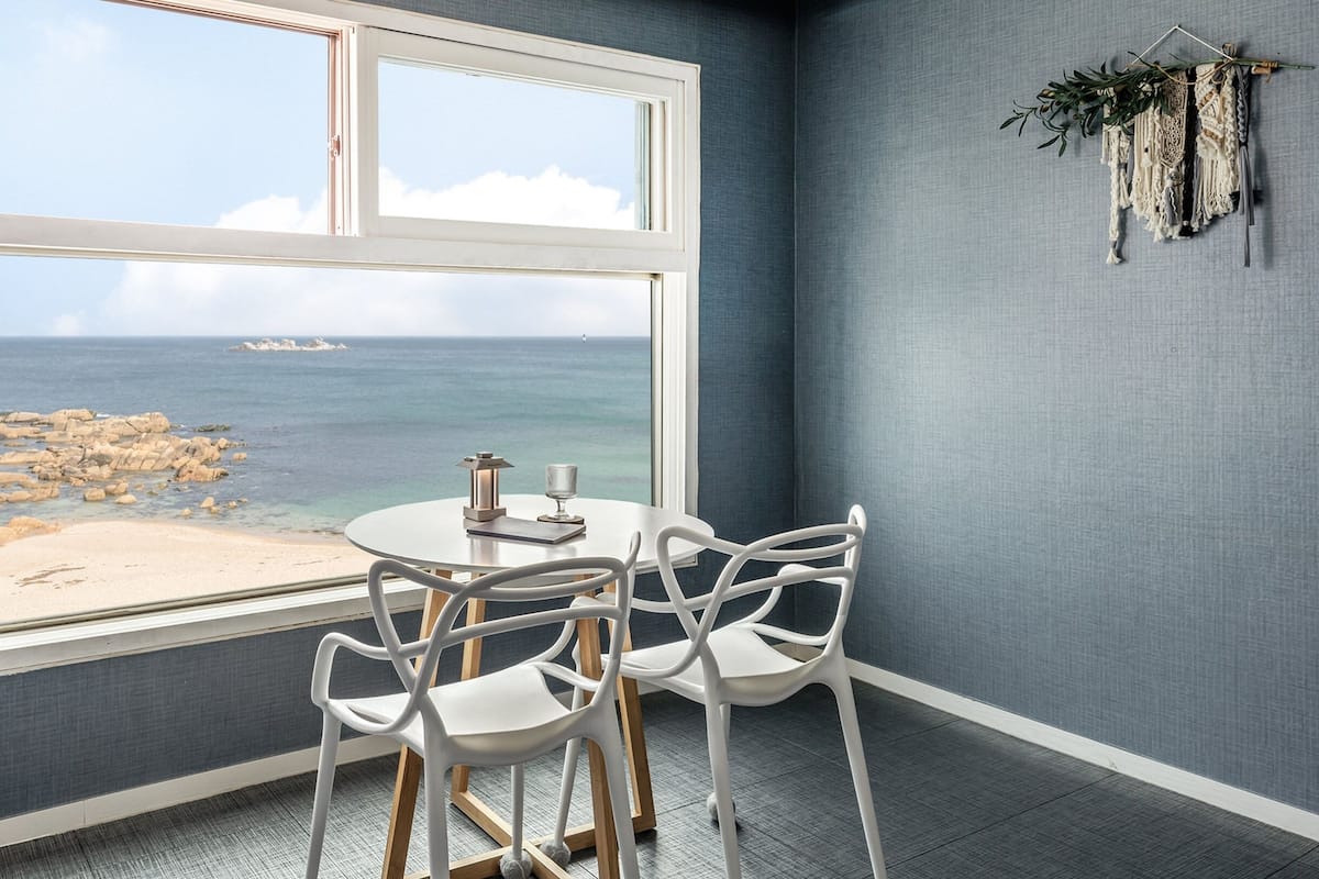 Una habitación con un amplio ventanal que da a una playa rocosa y al mar. Hay una mesa redonda con sillas y una lámpara pequeña sobre la mesa.