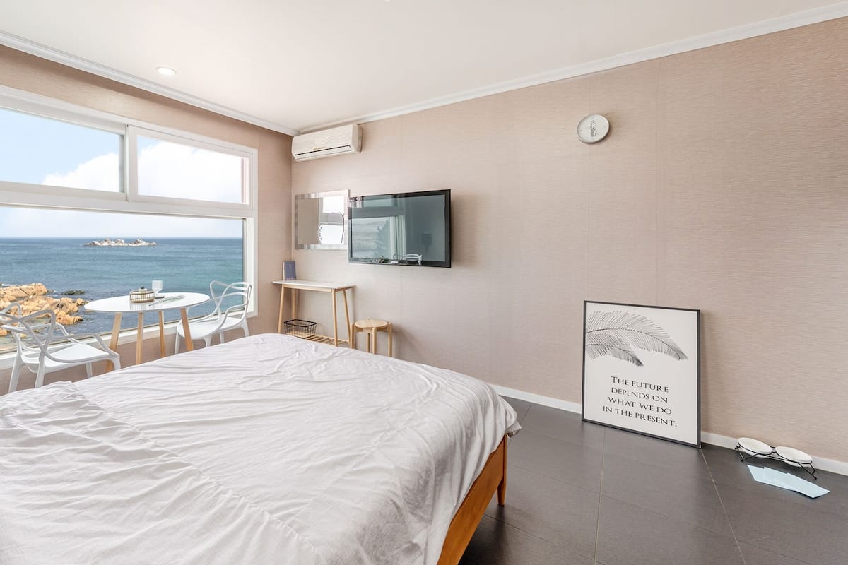 Un dormitorio con un ventanal grande con vista al mar, una cama, una pequeña mesa con sillas, un televisor montado en la pared y un cartel motivacional.