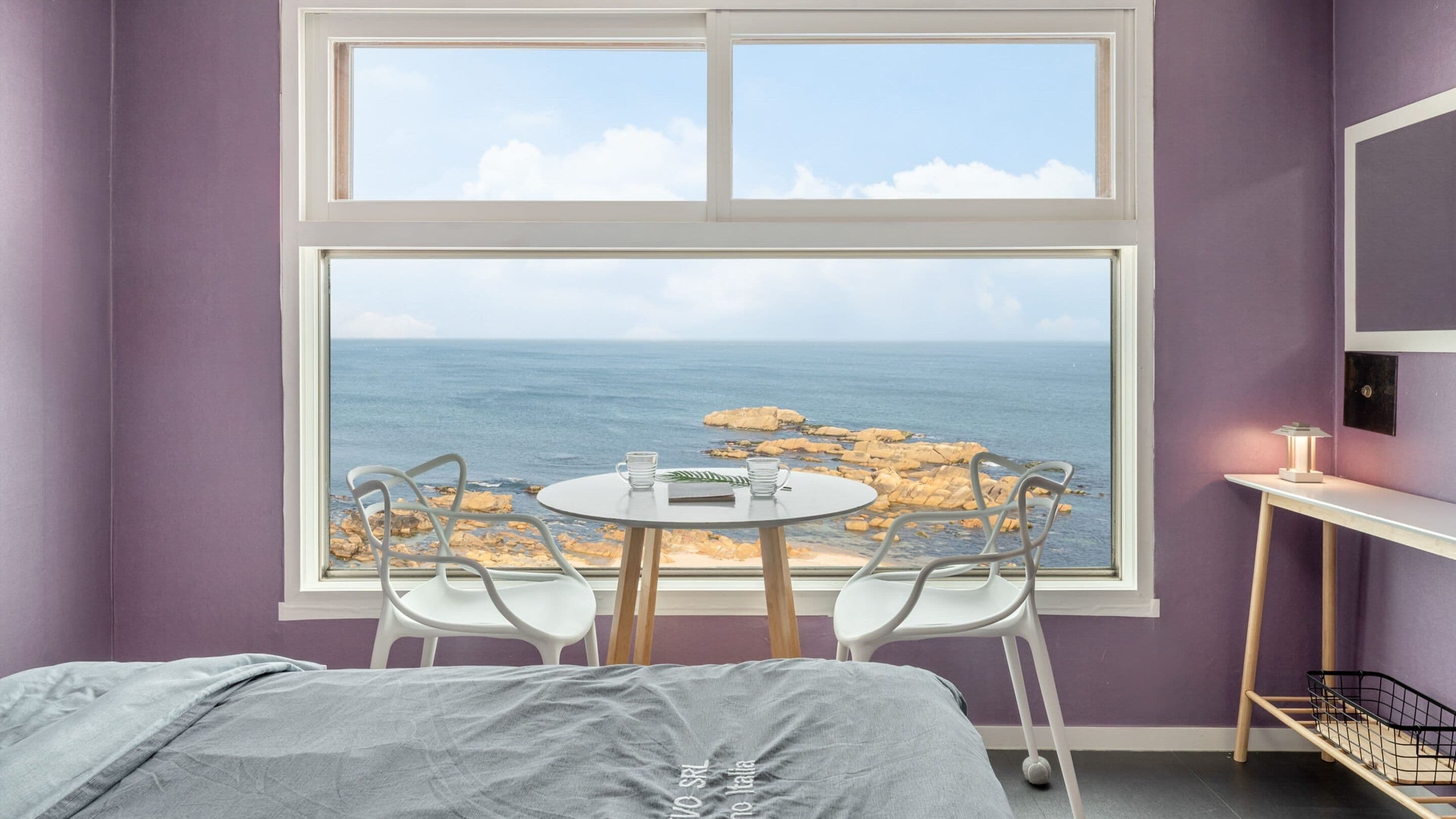Una habitación con un ventanal grande que da a una costa rocosa. Hay una cama con una manta gris, una mesita redonda con dos sillas y una mesita de noche con una lámpara.
