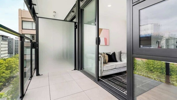 A modern balcony with a glass door, a sofa with cushions, and a city view. - Stylish Cozy Studio Apartment in CBD Near SkyTower (Auckland)