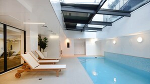 Een binnenzwembad met een glazen dak, ligbedden en een aangrenzende kamer met een plant.