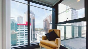 Een moderne kamer met een groot raam en uitzicht op de stad, een comfortabele stoel en een balkon met een glazen hekwerk.