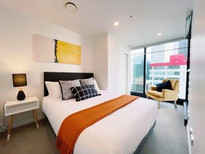 Quarto de hotel moderno com uma cama grande, uma mesa de cabeceira com abajur, uma cadeira e uma janela ampla com vista para a cidade.