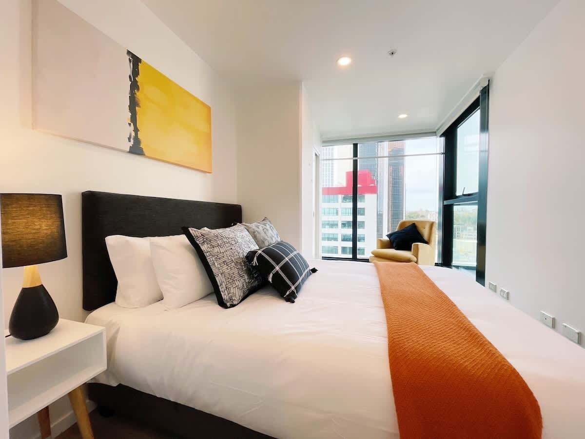 Ein modernes Hotelzimmer mit einem großen Bett, einem Nachttisch mit Lampe, einem Sessel und einem großen Fenster mit Blick auf die Stadt.