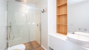 A modern bathroom with a glass shower enclosure, a white sink, and a wooden shelf above the mirror. - AG Villa Razo Deluxe, with swimming pool and beach 400m away (Razo Da Costa)