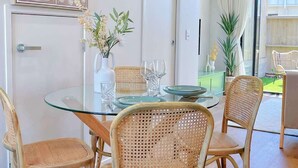 Un comedor con una mesa de vidrio rodeada de sillas de mimbre, un florero blanco con flores y una obra de arte abstracta colgada en la pared.