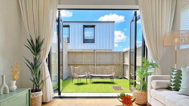 Una sala de estar moderna con un sofá, una mesa de centro con un florero, y vista a un patio trasero con un banco y un edificio pequeño.