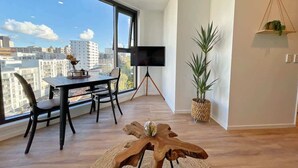 Un salon moderne doté d’une grande fenêtre, d’un téléviseur à écran plat posé sur un trépied, d’une table à manger avec des chaises et d’une table basse en bois.
