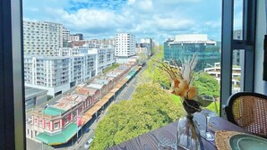 Une pièce dotée d’une grande fenêtre donnant sur un paysage urbain, avec notamment un bâtiment universitaire et une rue bordée d’immeubles et de verdure.