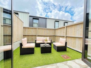 Un espace extérieur moderne, aménagé avec des meubles en osier et une table basse centrale, entouré d’une clôture en bois et surplombé par un ciel dégagé.